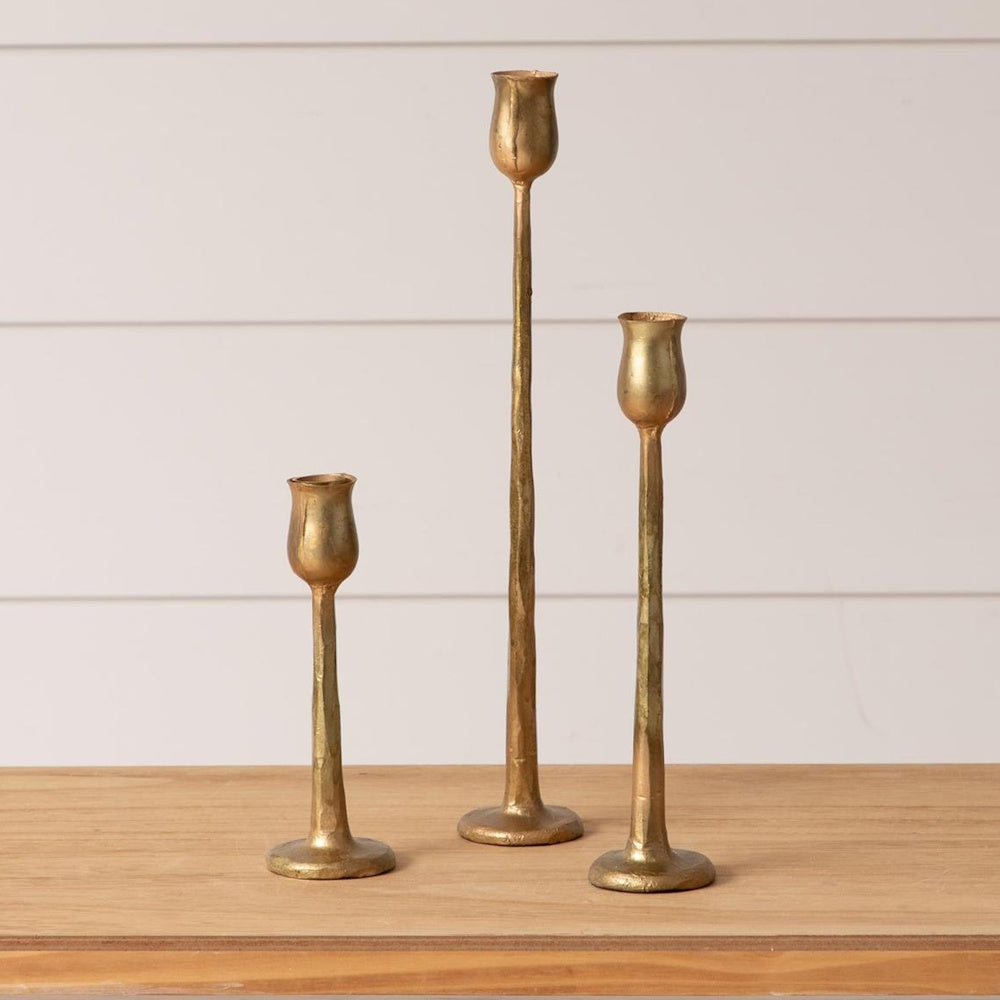 Set of 3 slender gold metal taper candle holders in varying heights with a rustic, hand-hammered finish on a wood surface.