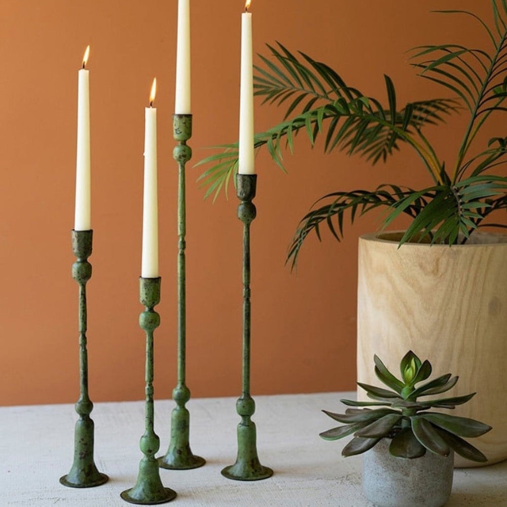 Four slender, green patina taper candle holders with lit white candles, standing next to potted plants.