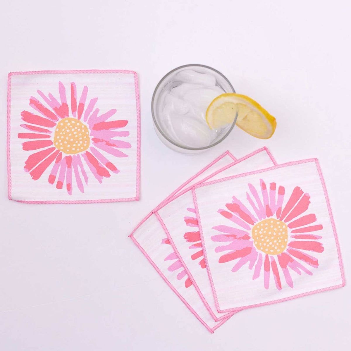 Pink daisy cocktail napkins next to an iced drink with a lemon slice on a white surface.