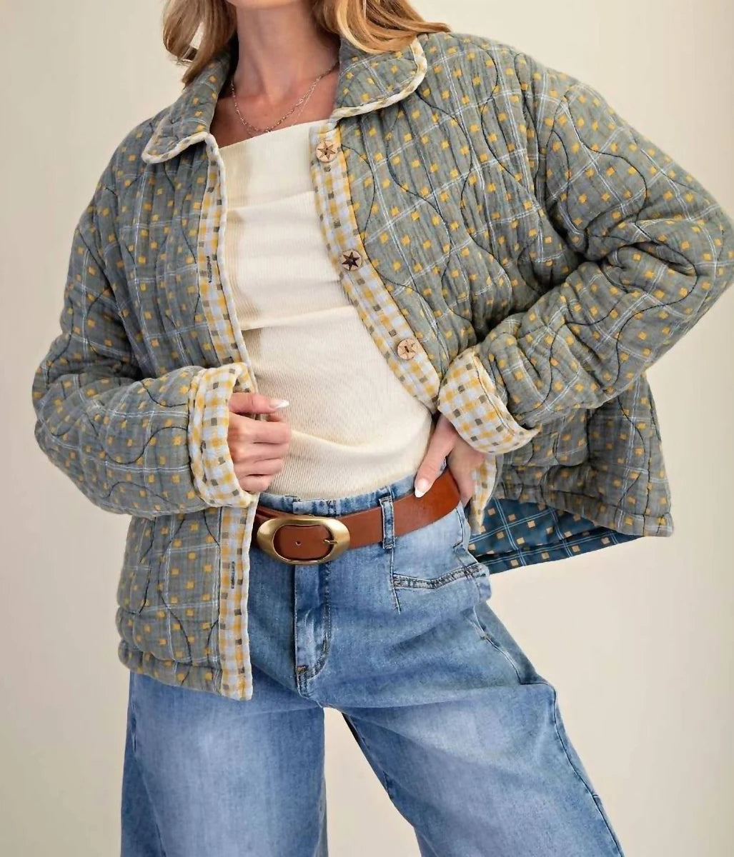 A woman models a quilted jacket with a muted blue-gray and mustard yellow gingham pattern, styled with a cream ribbed top, blue jeans, and a brown belt.