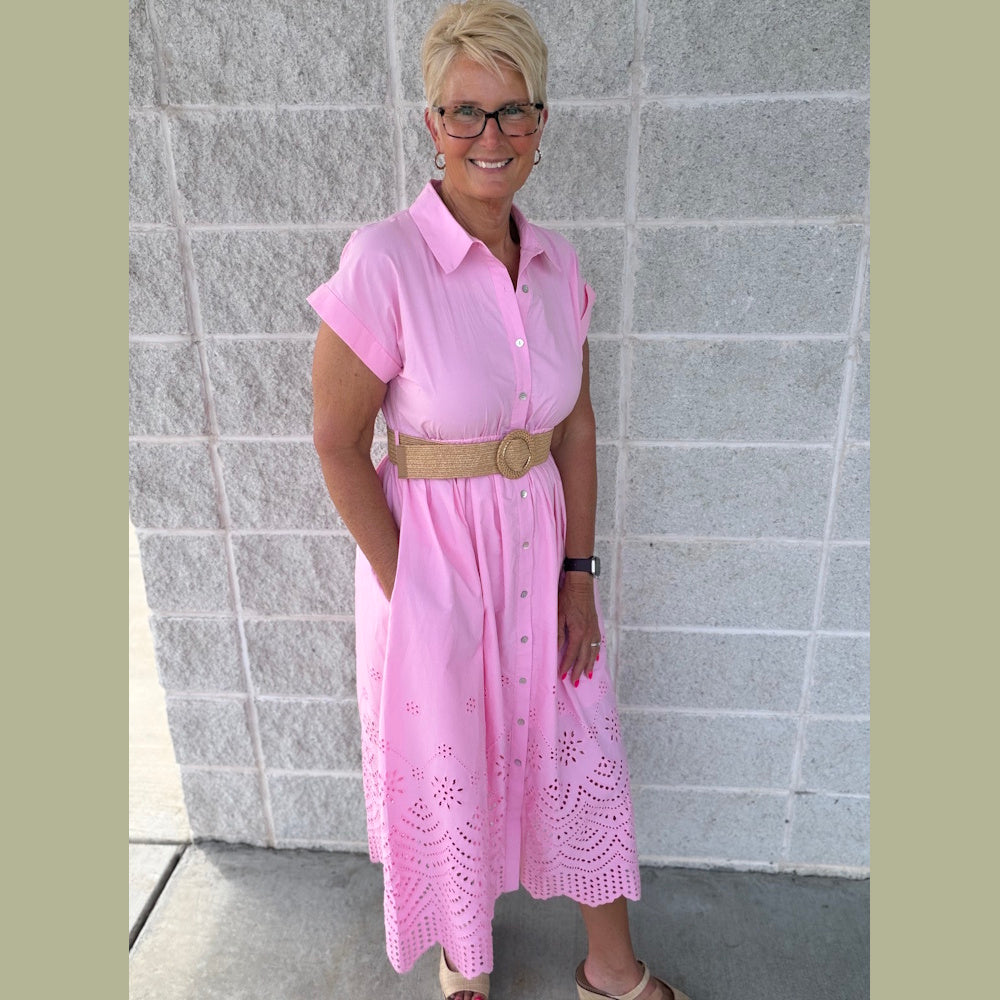A woman in a pink short-cuffed sleeve midi dress with eyelet detailing and a woven belt stands against a wall.