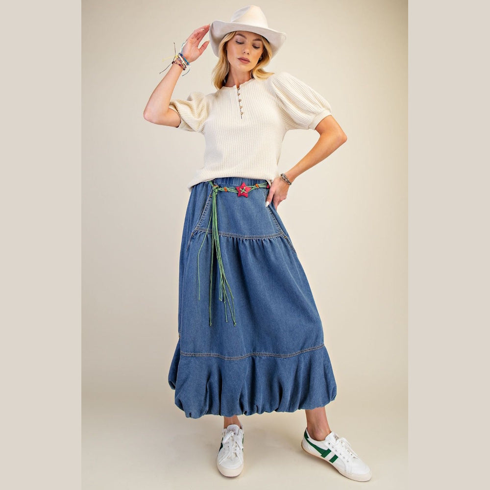 Woman in a cream waffle knit puff sleeve top and long denim tiered skirt with a white wide-brim hat.