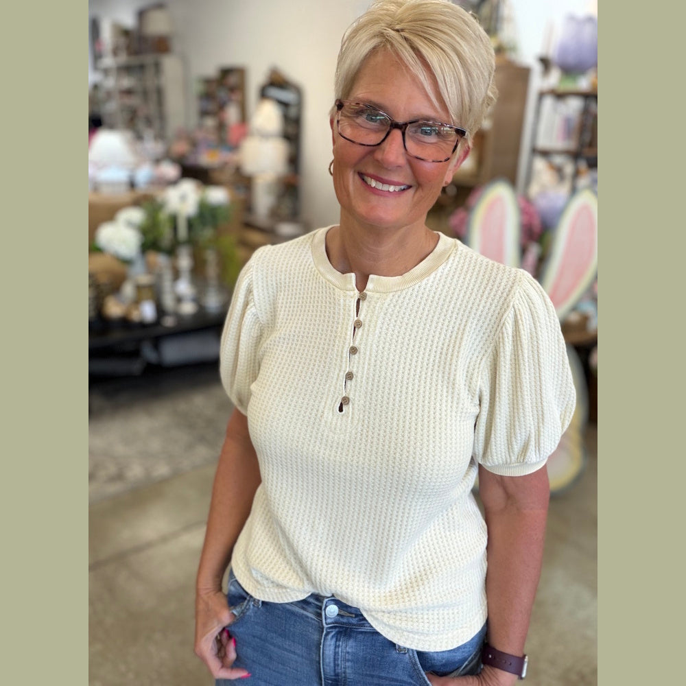 Smiling woman in glasses wearing a cream knit top and jeans in a home decor shop.