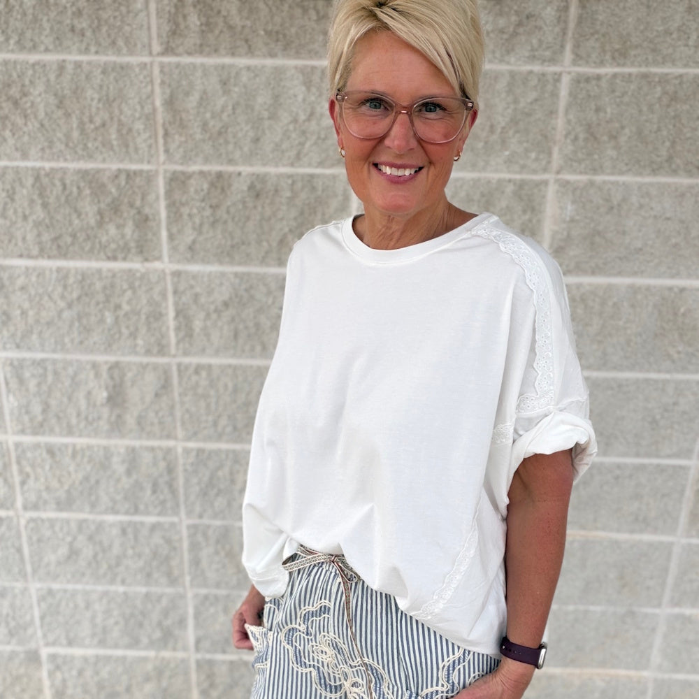 Woman in a white short-sleeve top with lace detail and striped pants smiling in front of a gray brick wall.