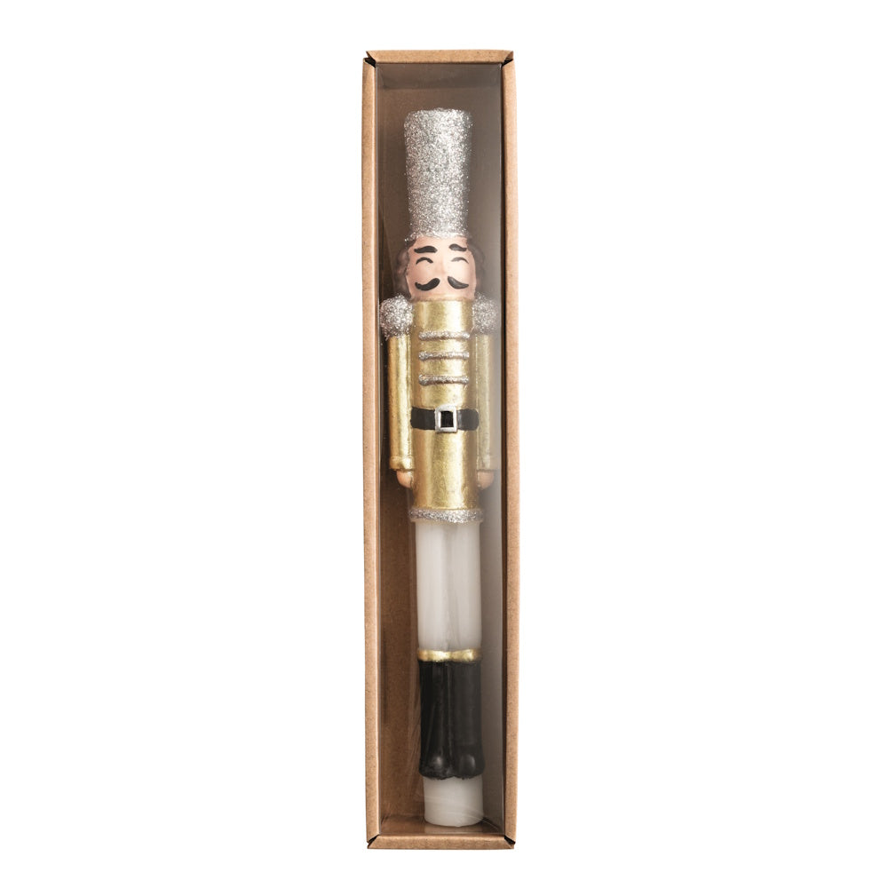 Front view of a gold and white wax nutcracker taper candle with silver glitter details in a boxed package.