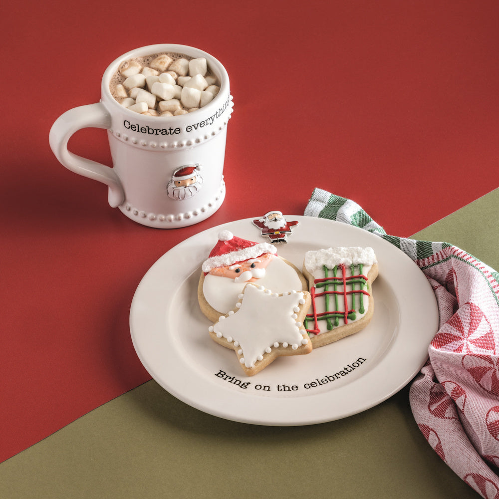 Christmas mug of hot cocoa with marshmallows and Santa cookies on a "Bring on the celebration" plate.