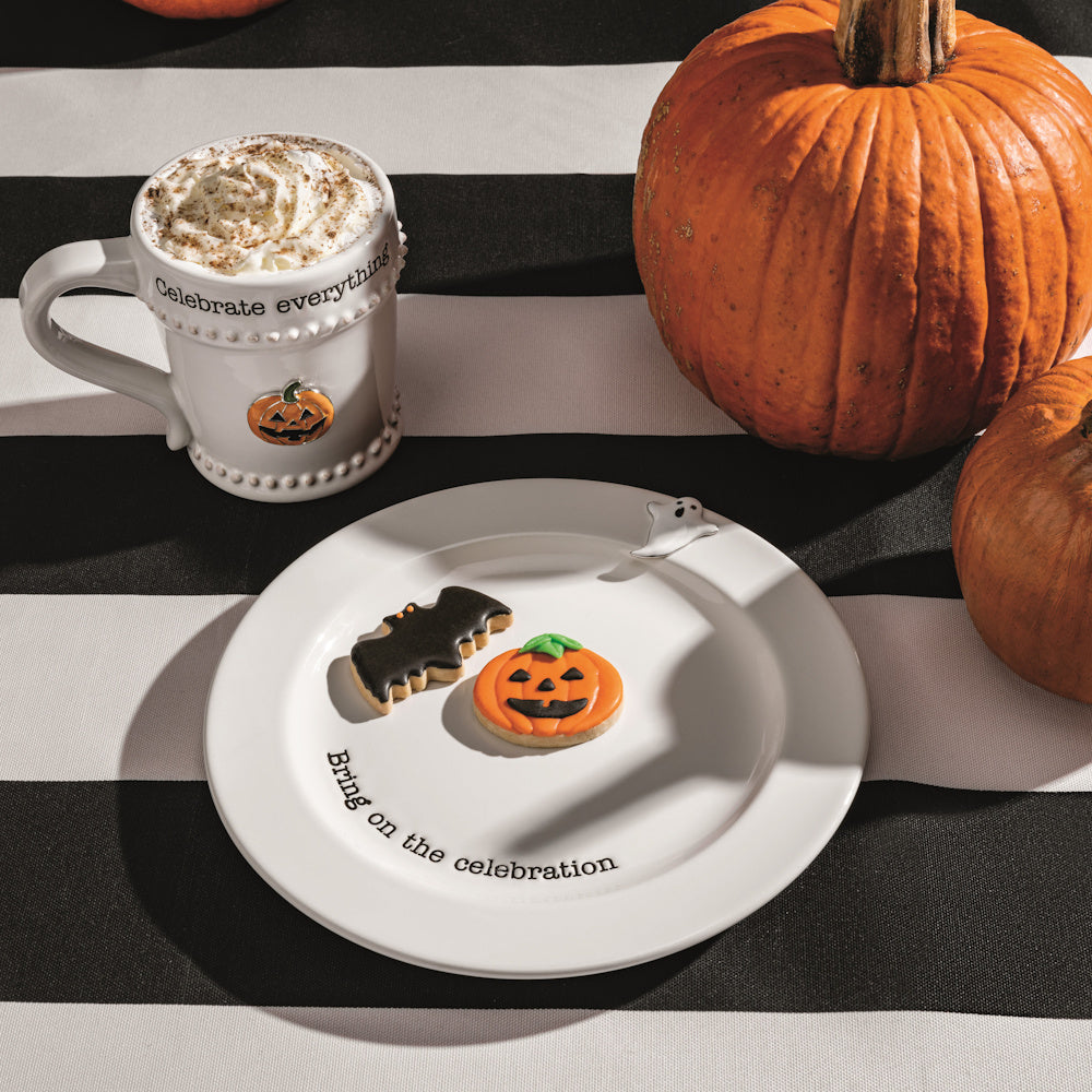 Halloween-themed plate with bat and pumpkin cookies next to a festive mug and pumpkins on a striped tablecloth.
