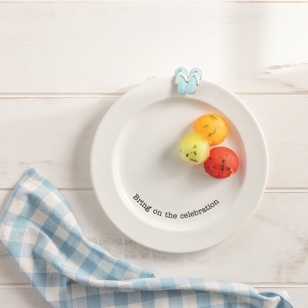 White ceramic celebration plate with flip-flop charm, melon balls, and blue gingham napkin on a wood table.