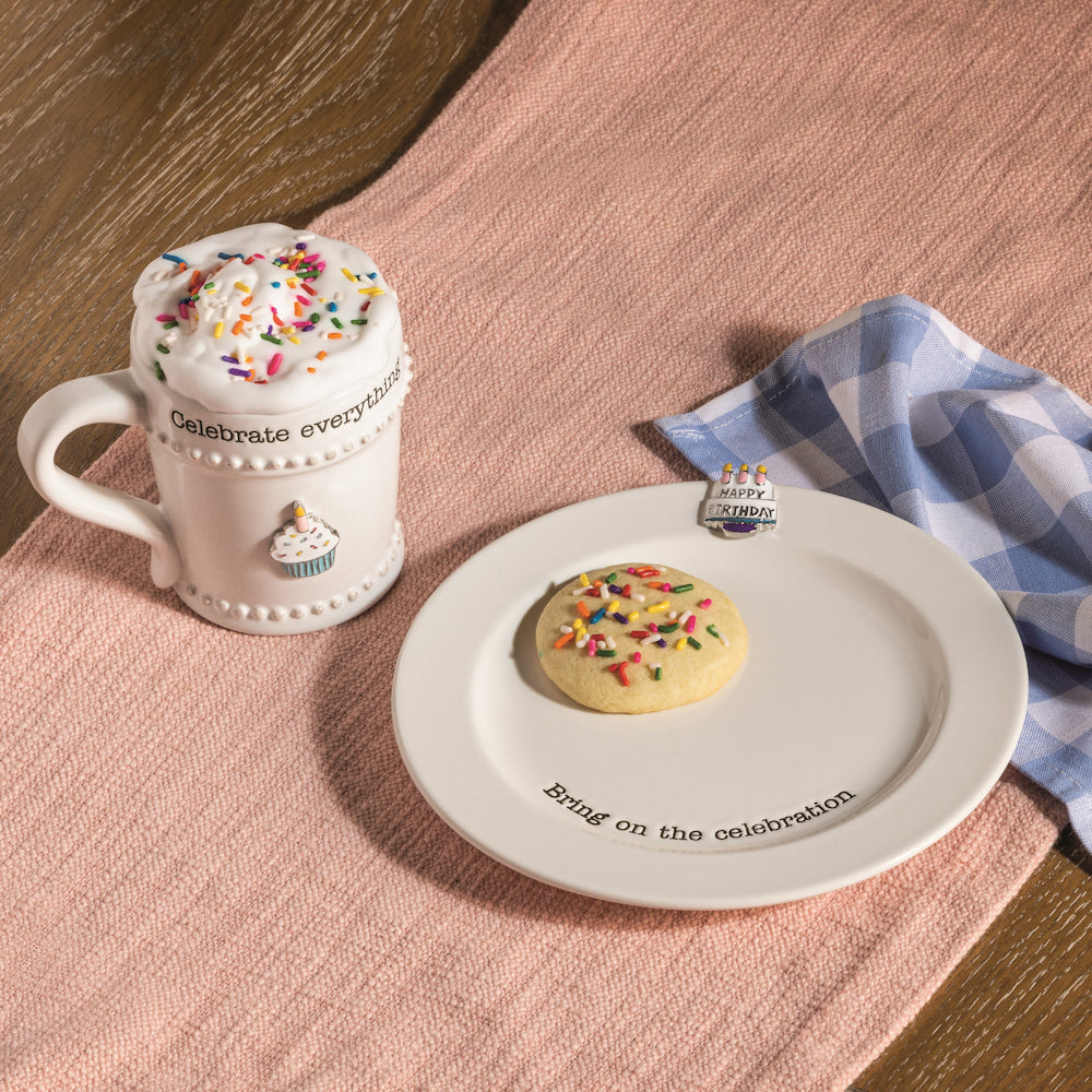 White ceramic mug and cookie plate with sprinkles and birthday charms on a pink runner.