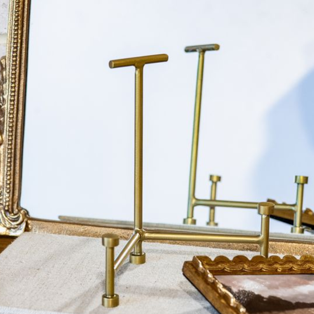 Antique brass display easel with a T-bar top and round-footed legs set near framed art.