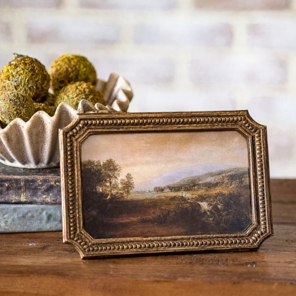 Framed landscape art in a beaded antique gold frame, set on a wooden table next to stacked books.