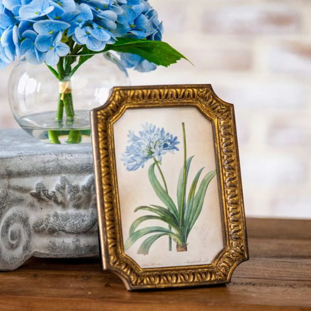 Blue flower art print in an ornate gold frame set on a wooden surface next to blue hydrangeas.