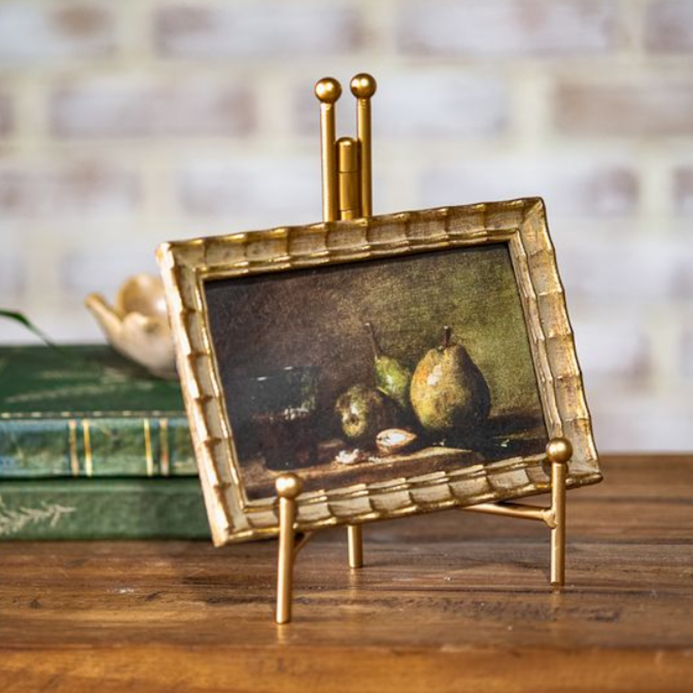 Framed fruit still life painting in a scalloped gold frame on a metal easel next to stacked books.