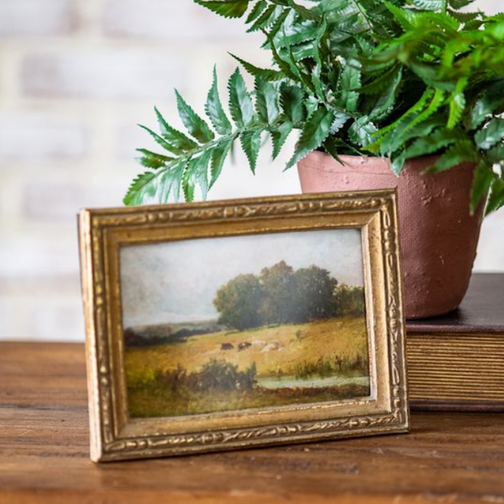Small framed landscape painting in an antique gold frame on a wooden surface next to a potted plant.