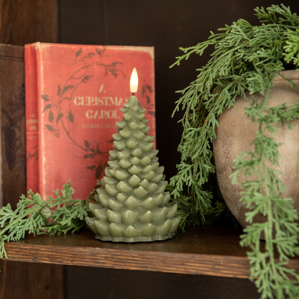 A lit tree-shaped green LED candle on a shelf next to a red Christmas book and greenery.