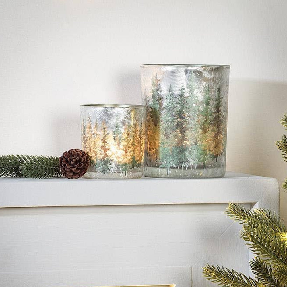 Two winter forest glass candle holders displayed on a white mantel with pine needles and a pinecone.