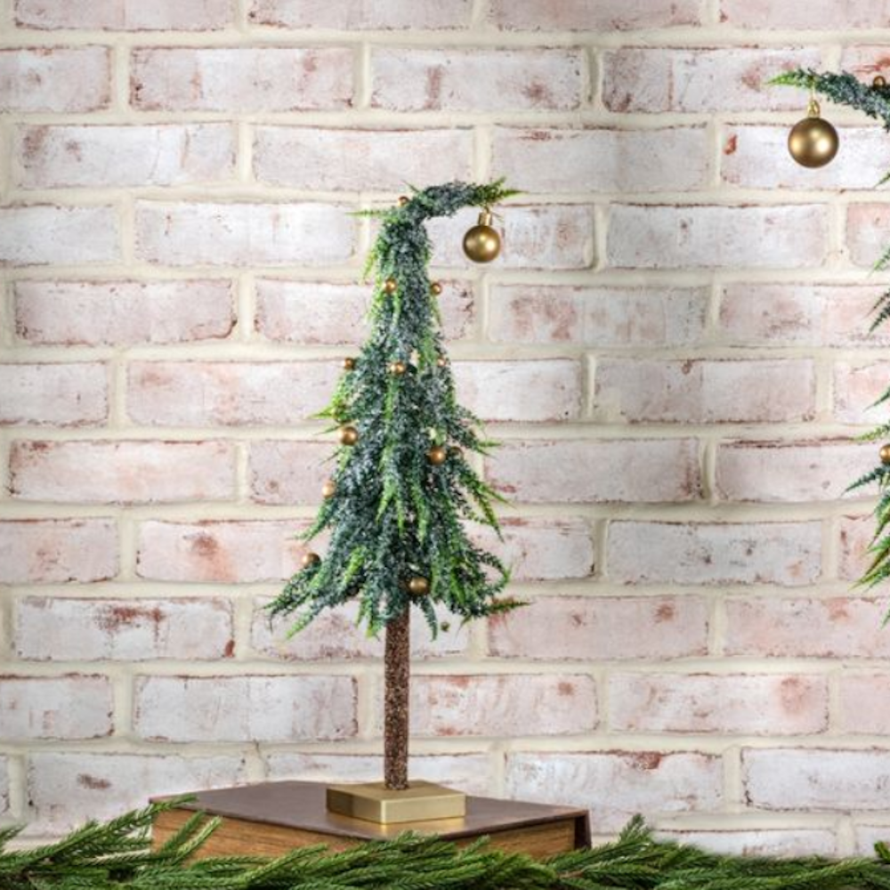 Small frosted tabletop evergreen tree with gold ornament on a stand, placed in front of a brick wall.