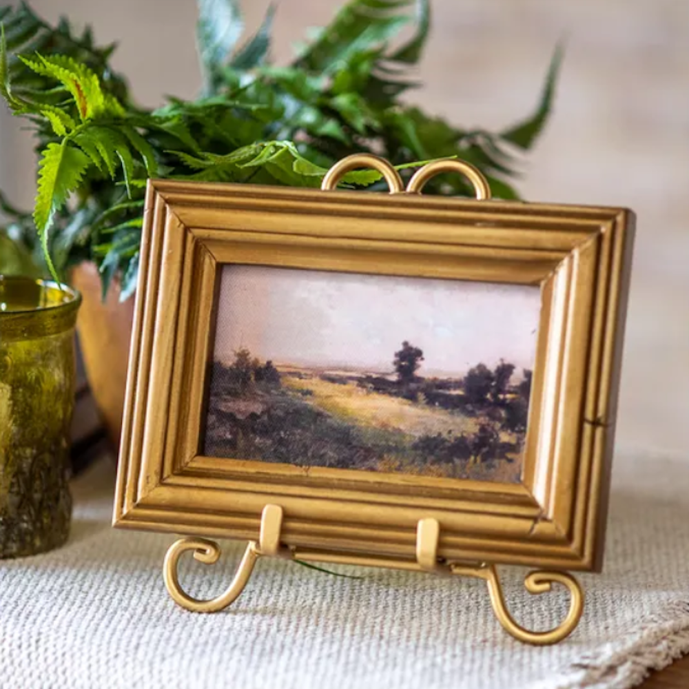 Framed mini landscape painting depicting a countryside scene resting on a gold stand.