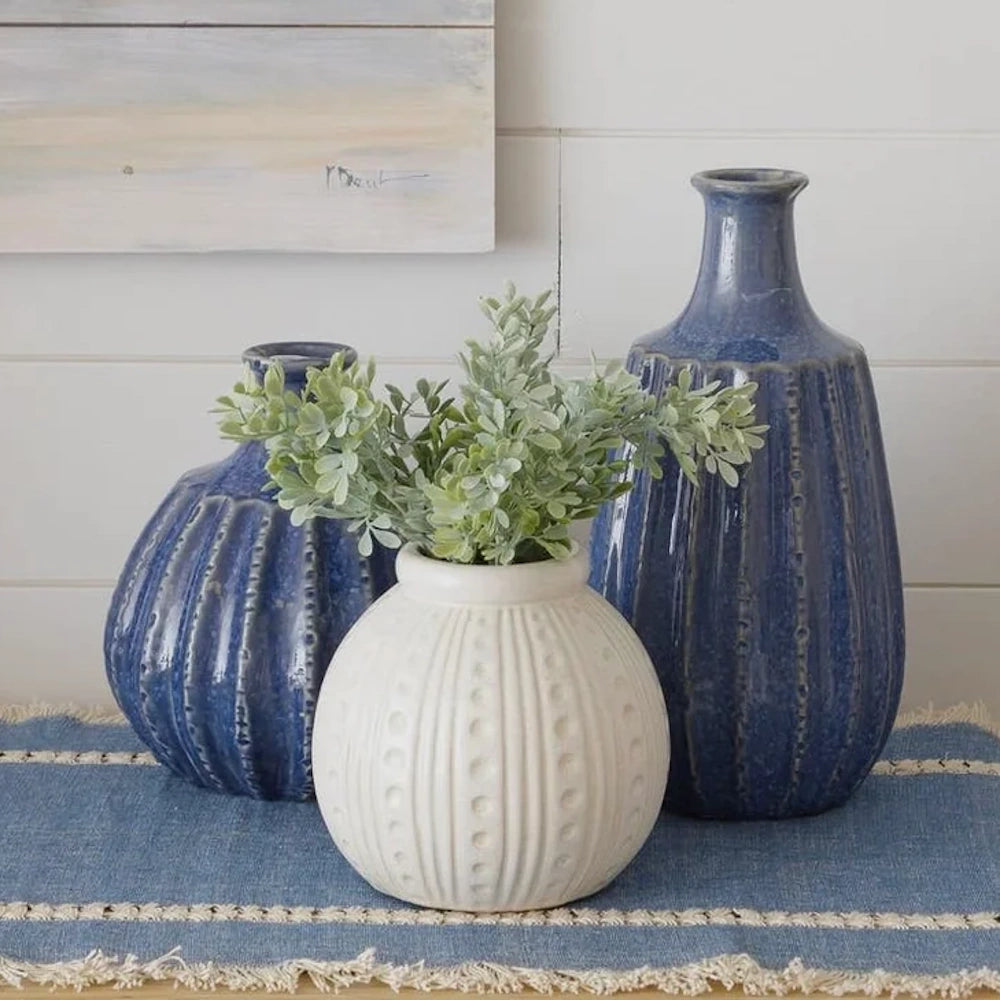Three textured ceramic vases, two blue and one white with green foliage, on a blue runner.