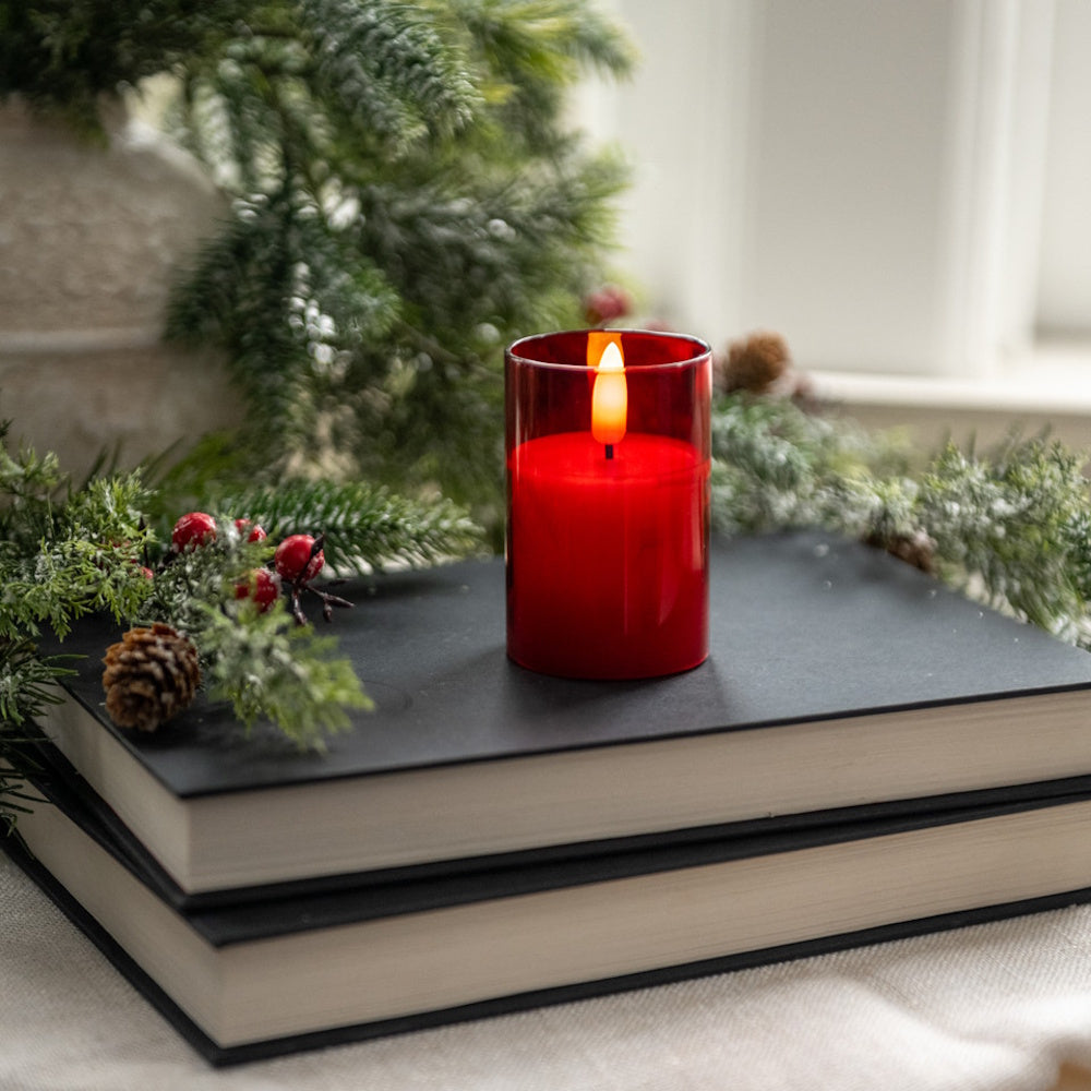 A small glowing red glass candle resting on stacked black books with pine greenery and red berries.