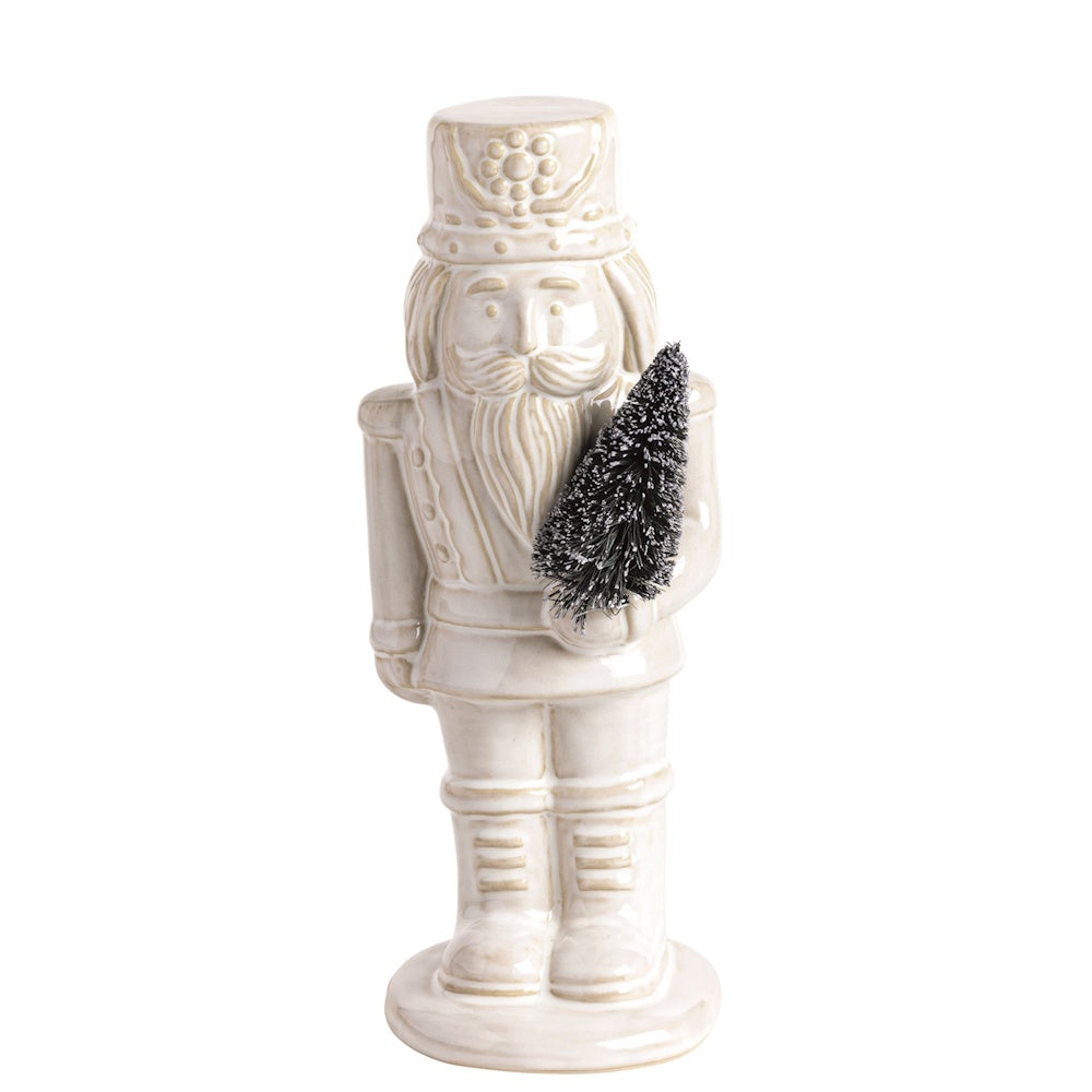 White ceramic nutcracker figurine holding a small frosted miniature pine tree.