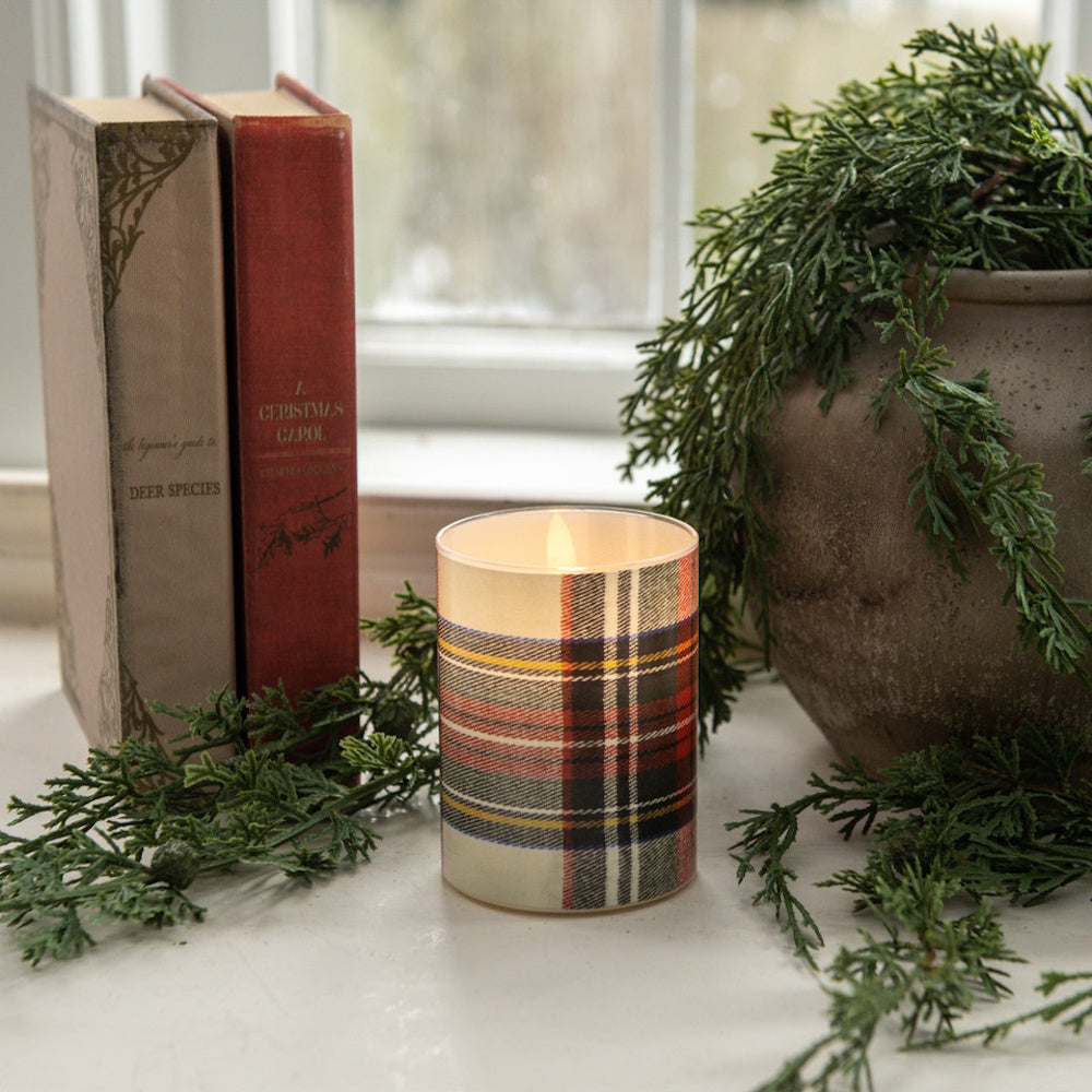 A small glowing flameless candle wrapped in a white tartan plaid pattern by books and greenery.