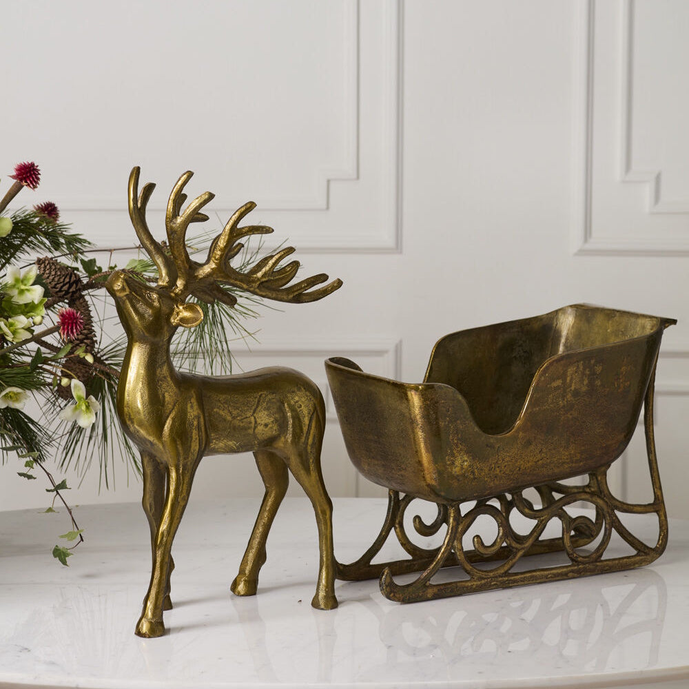 Gold reindeer figurine standing in front of a matching brass sleigh on a marble surface.