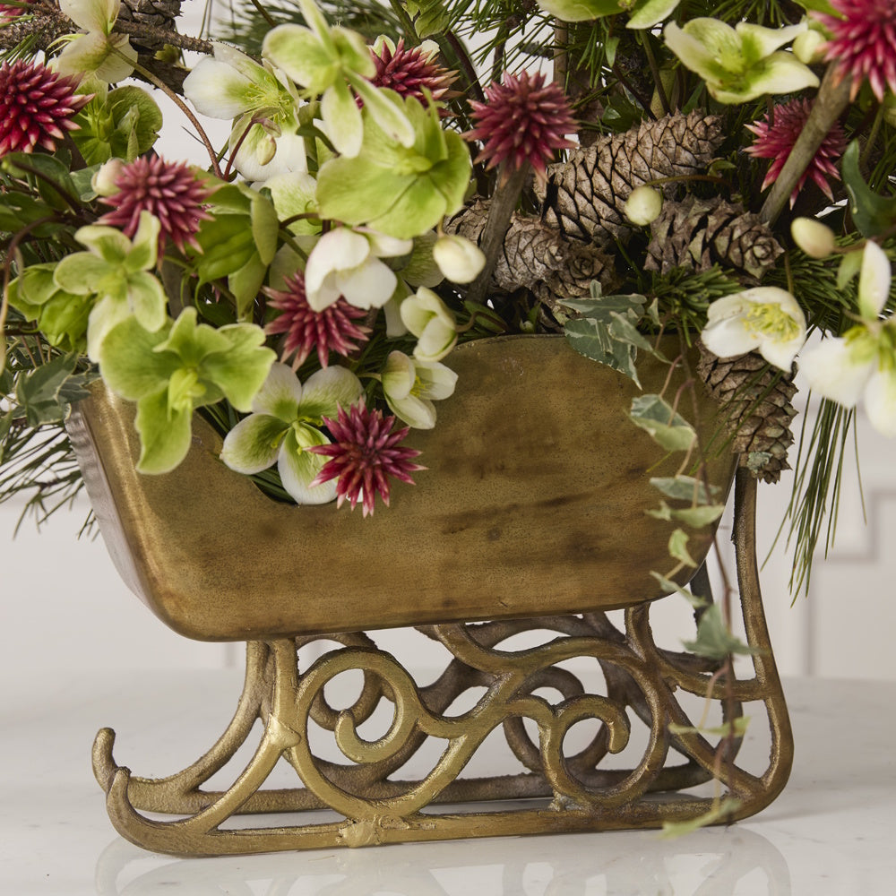 Vintage gold sleigh planter filled with green and burgundy flowers, pinecones, and greenery.