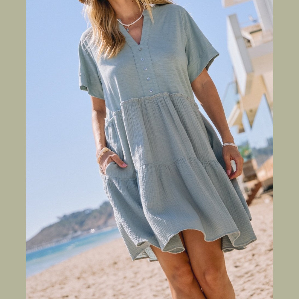 Woman standing on a beach wearing a short-sleeved, tiered sage green mini dress with a buttoned neckline.