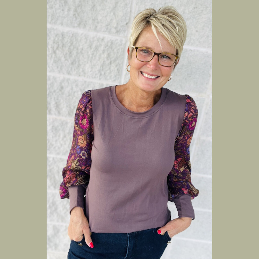 Woman with short blonde hair and glasses wearing a solid purple top with floral sleeves.