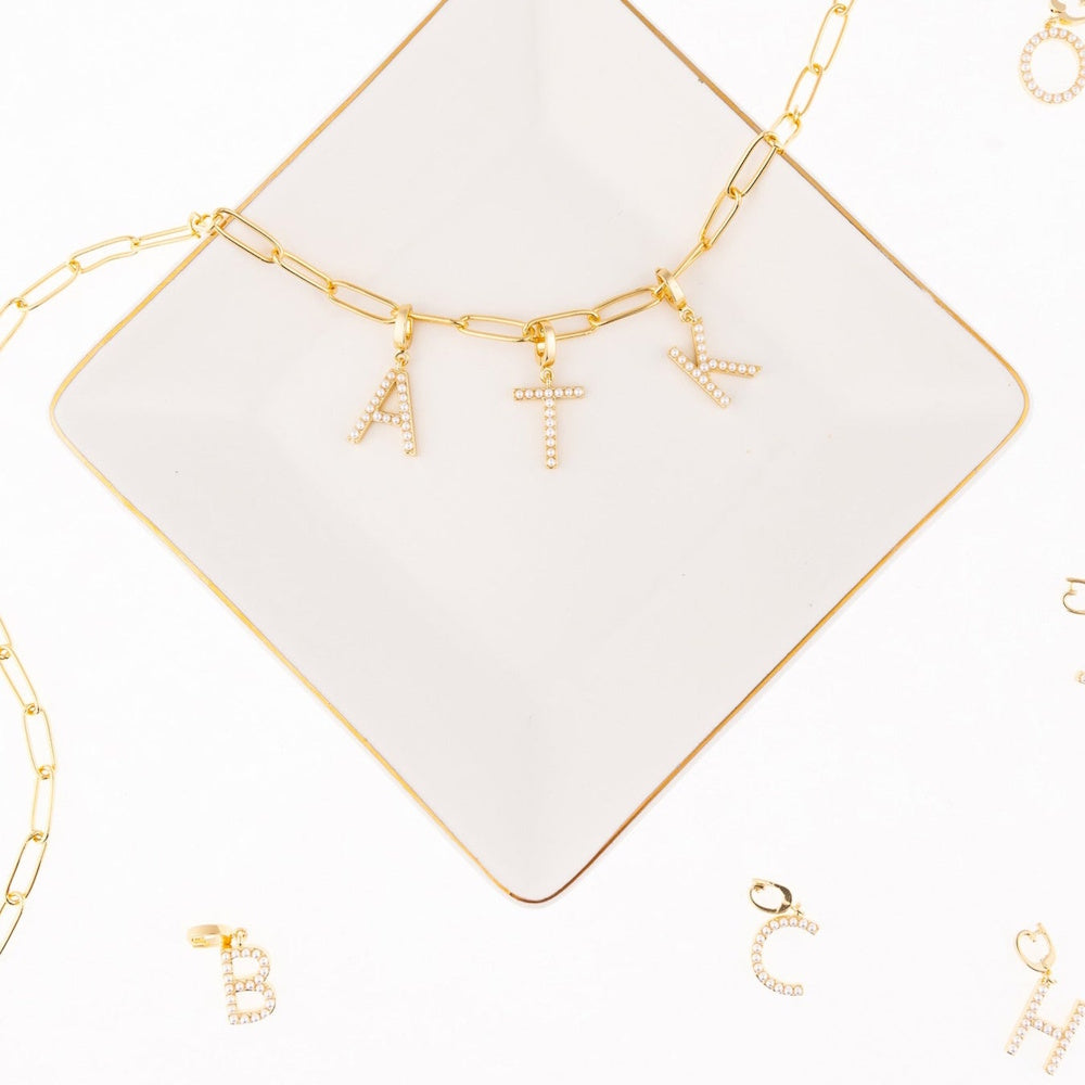 Gold link chain necklace with faux pearl initial charms A, T, and K resting on a square dish.