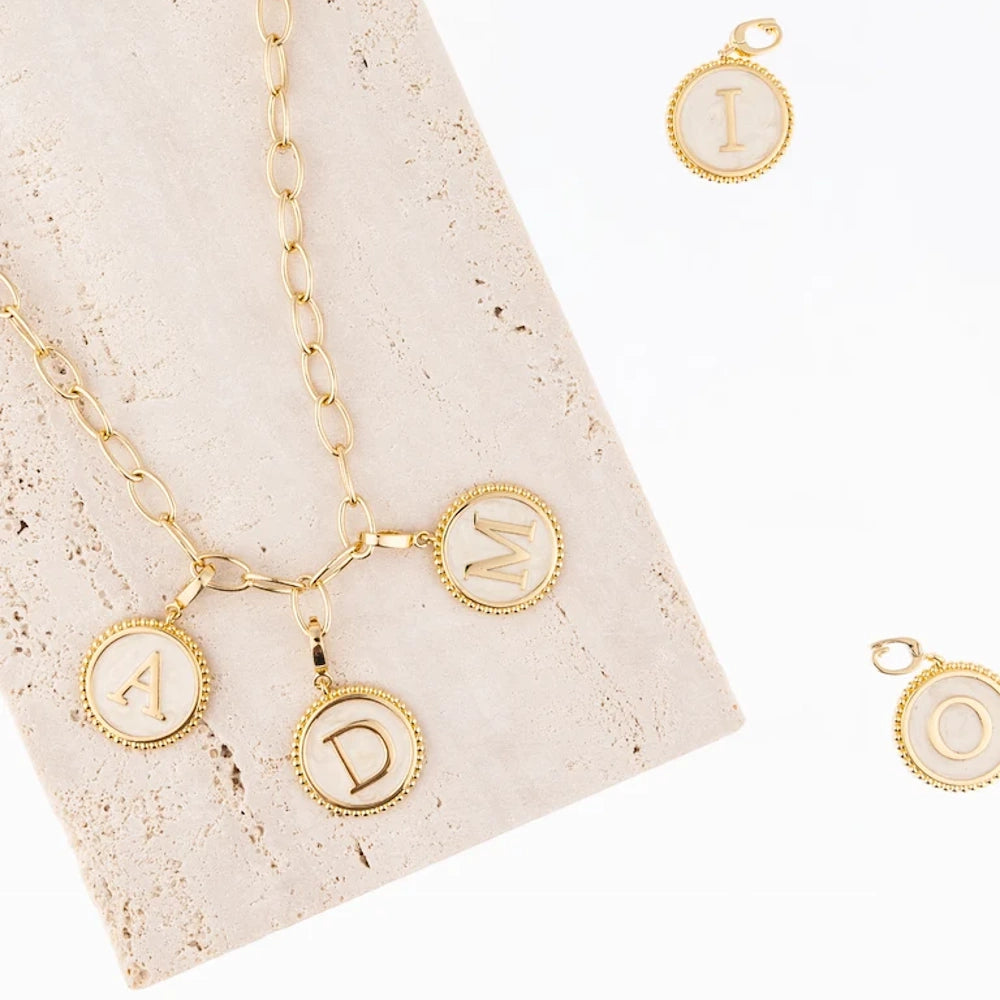 Gold link chain necklace with faux mother-of-pearl initial charms on a stone block alongside extra charms.