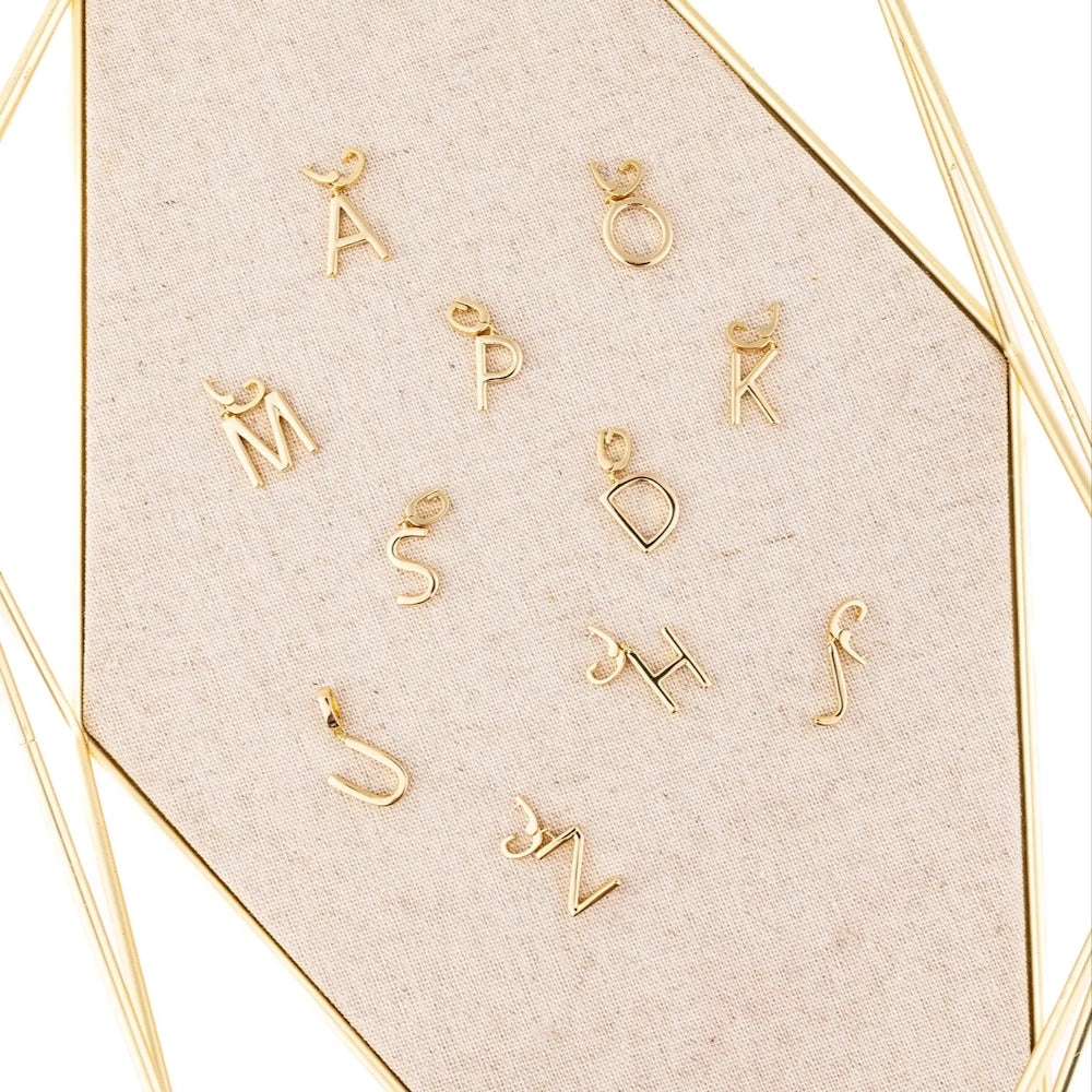 Polished gold initial letter charms arranged on a textured beige fabric display tray with gold geometric trim.