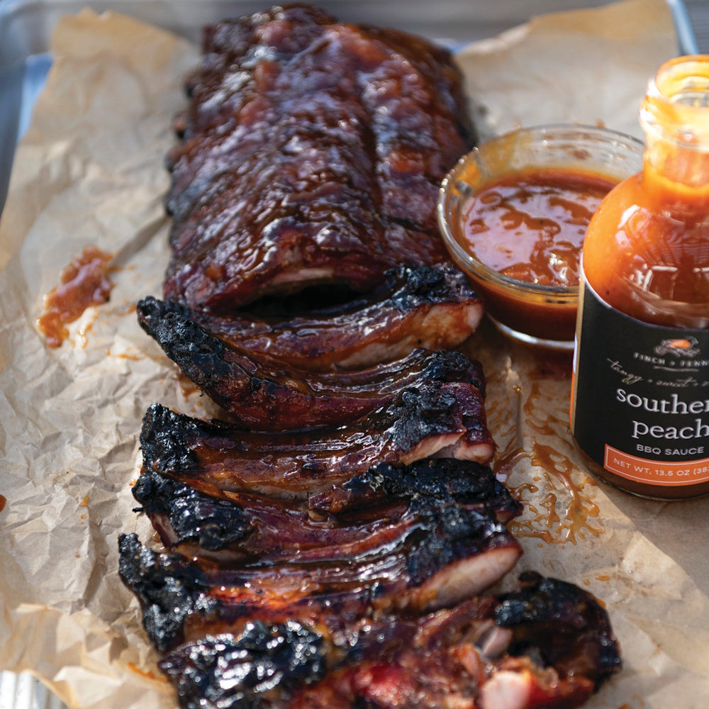 Glazed, sliced BBQ ribs on parchment paper next to a bottle of Finch + Fennel Southern Peach BBQ sauce.