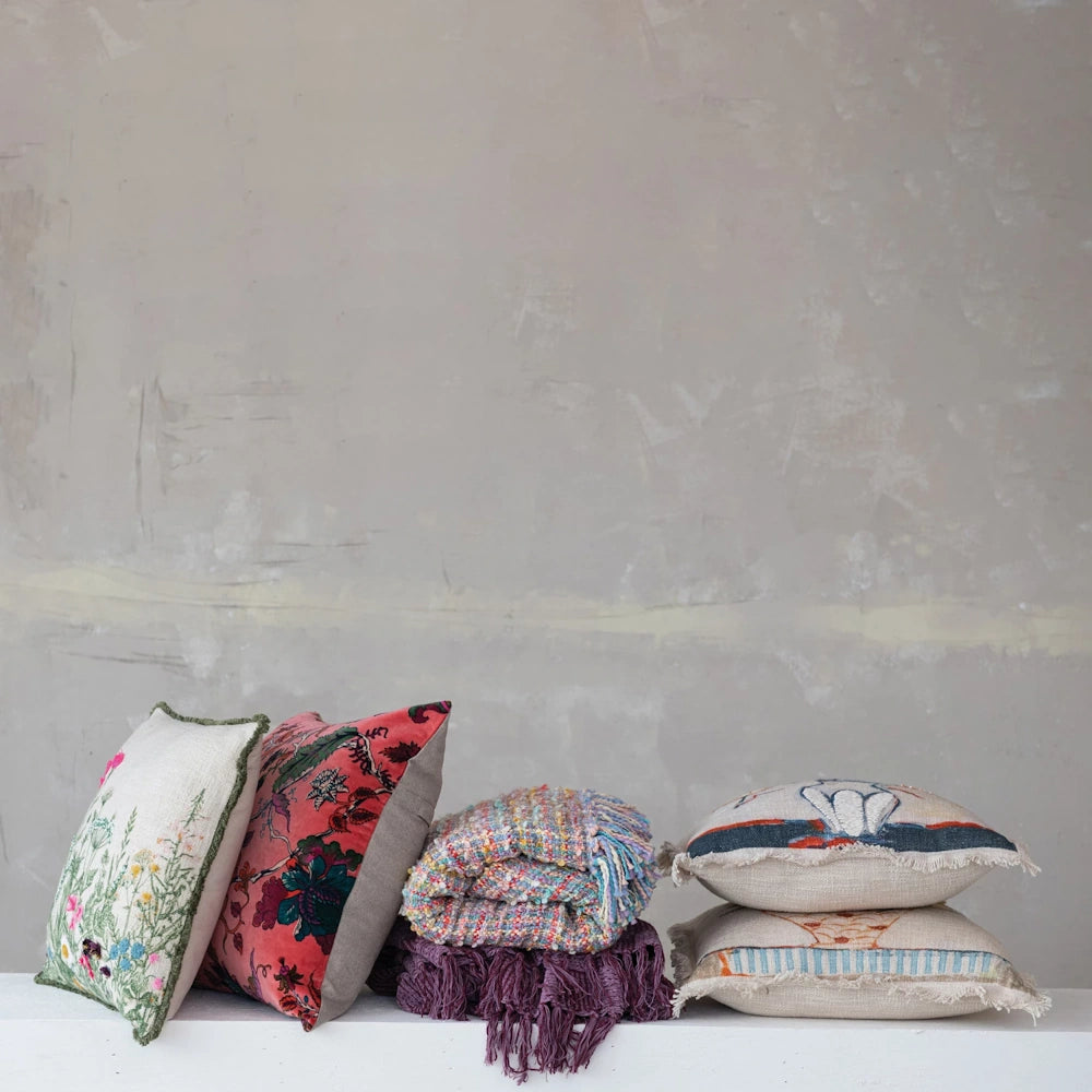 Colorful embroidered throw pillows and textured blankets stacked against a neutral, rustic plaster wall.