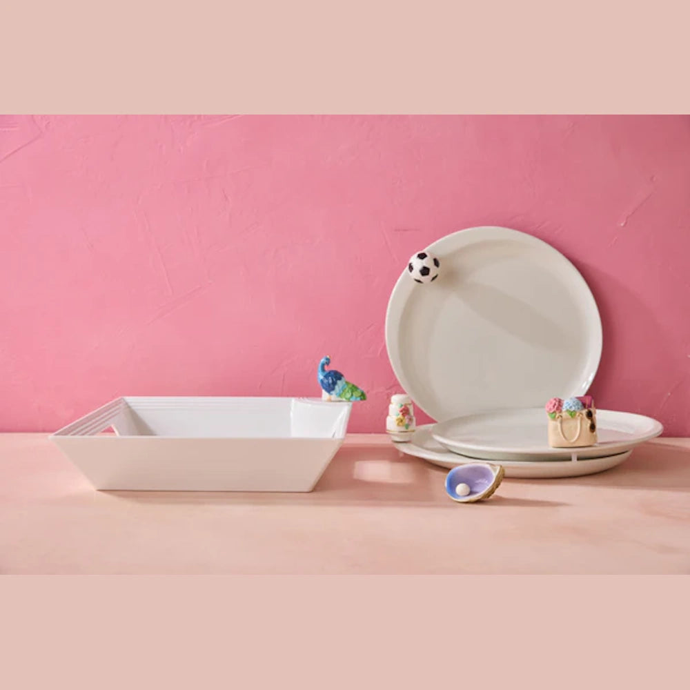 A still life of square and stacked round white ceramic serving dishes and decorative trinkets against a textured pink wall.