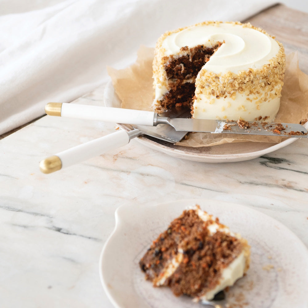 Lifestyle view of the stainless steel cake server and knife set with white and gold handles used to slice a frosted layer cake on a table.