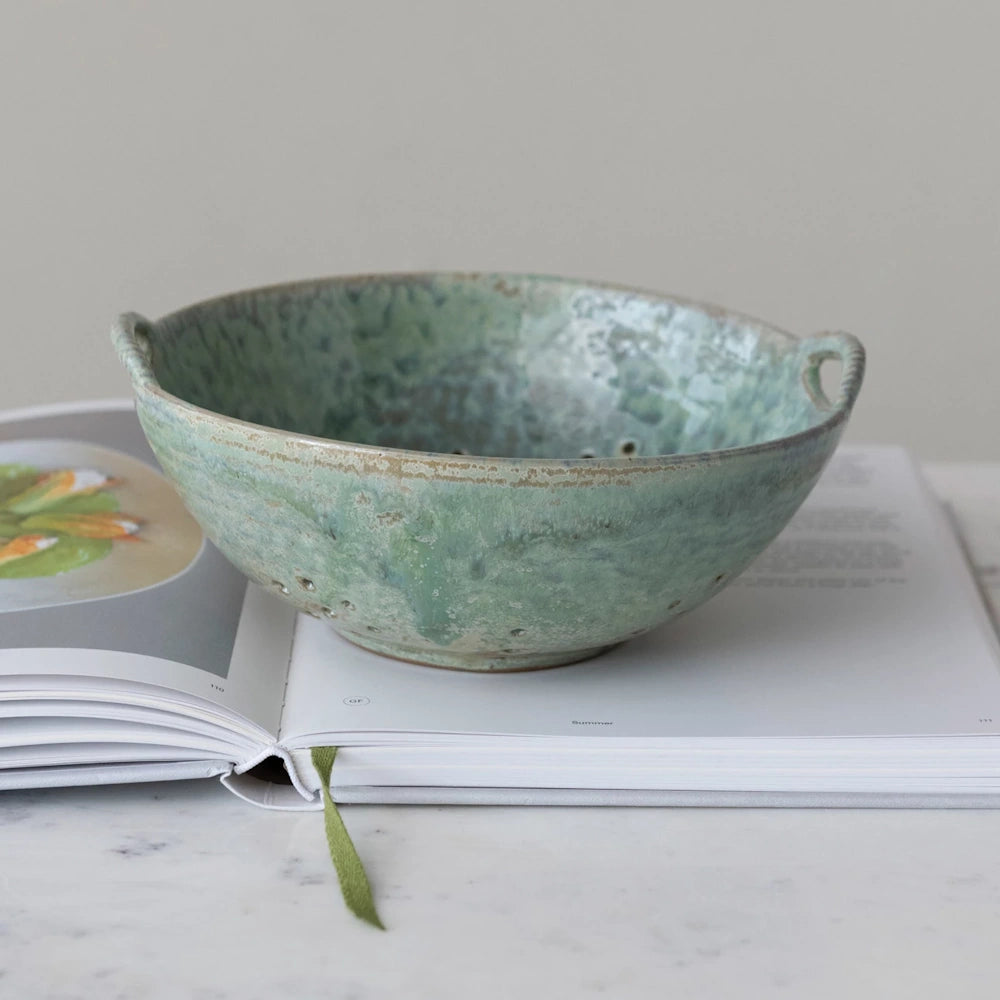 Handmade green stoneware berry bowl with handles and drainage holes, resting on an open cookbook.