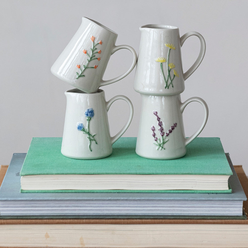 Four stoneware creamers with embossed botanical designs stacked on top of books.
