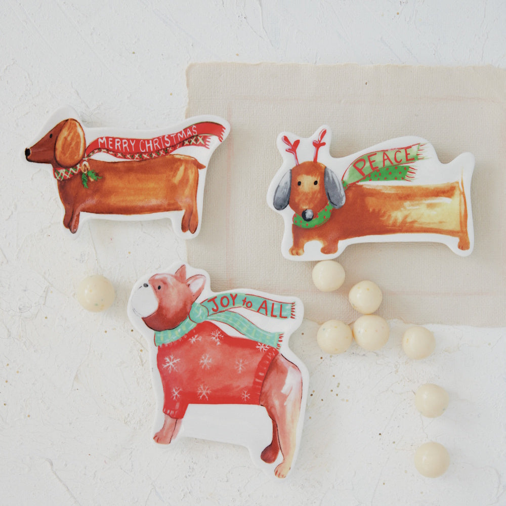 Three stoneware Christmas dog dishes arranged on a light surface with small round candies.