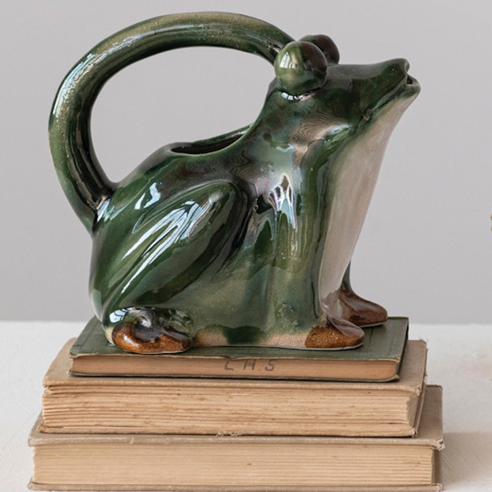 A shiny green ceramic frog pitcher with a curved handle, sitting on a stack of three vintage books.