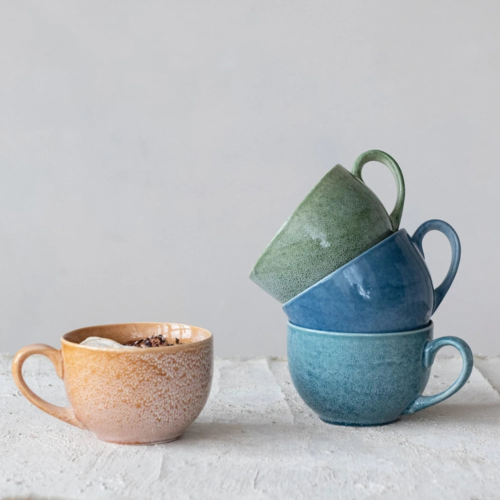 Stacked green and blue stoneware mugs next to an orange ceramic coffee cup on a textured white surface.