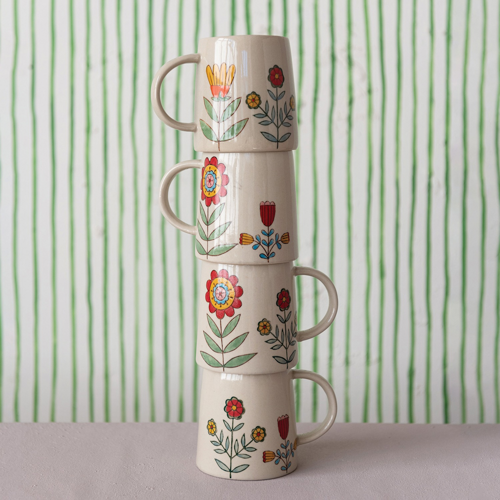 Stacked stoneware mugs with hand-painted floral patterns against a green striped background.