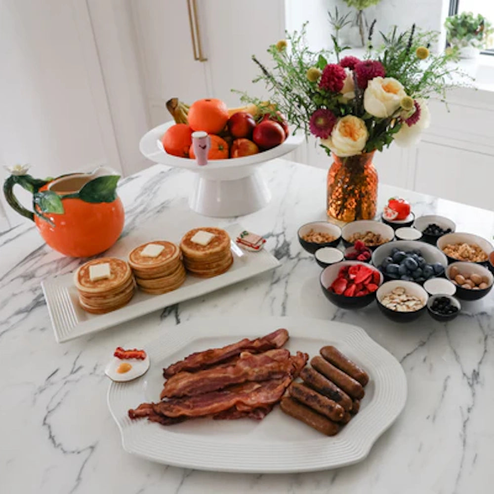 Breakfast spread with pancakes, bacon, sausage, and fruit toppings on white stoneware platters over a marble table.
