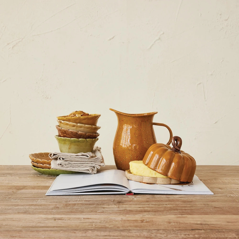 Stoneware pumpkin-shaped butter dish with butter inside on an open book, next to a pitcher and stacked bowls.