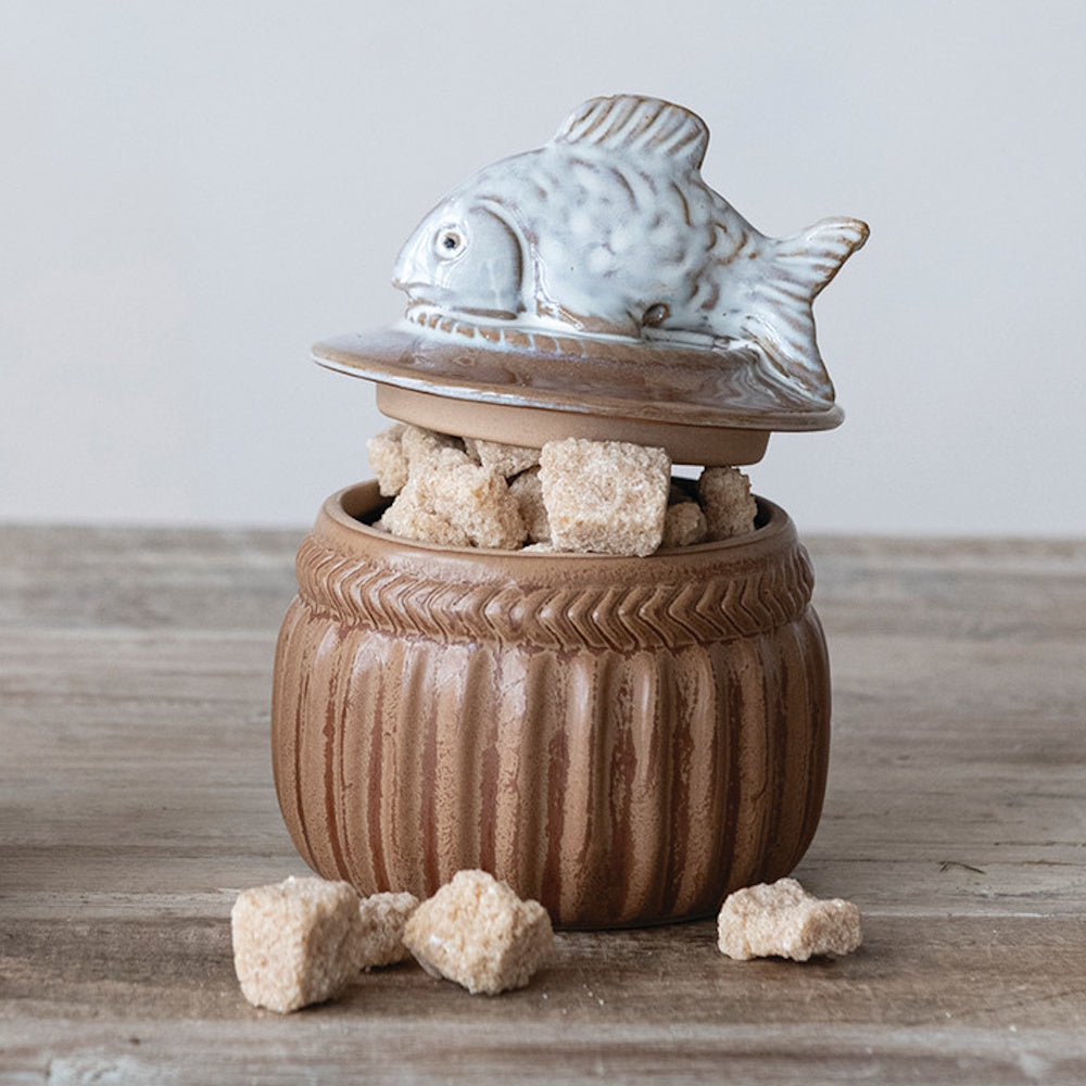 Brown stoneware salt cellar with a decorative fish lid, filled with brown sugar cubes on a rustic wooden surface.