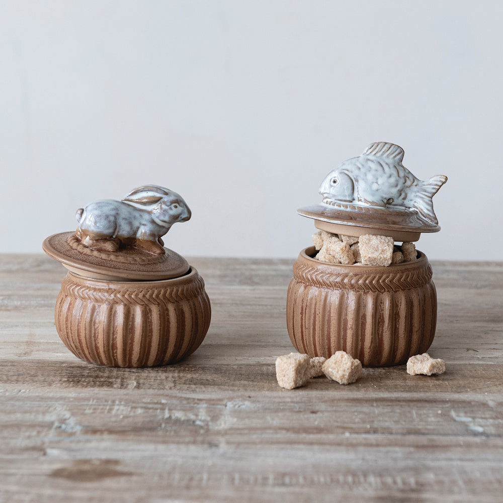 Two brown stoneware cellars with lids featuring white rabbit and fish figurines, filled with brown sugar cubes.