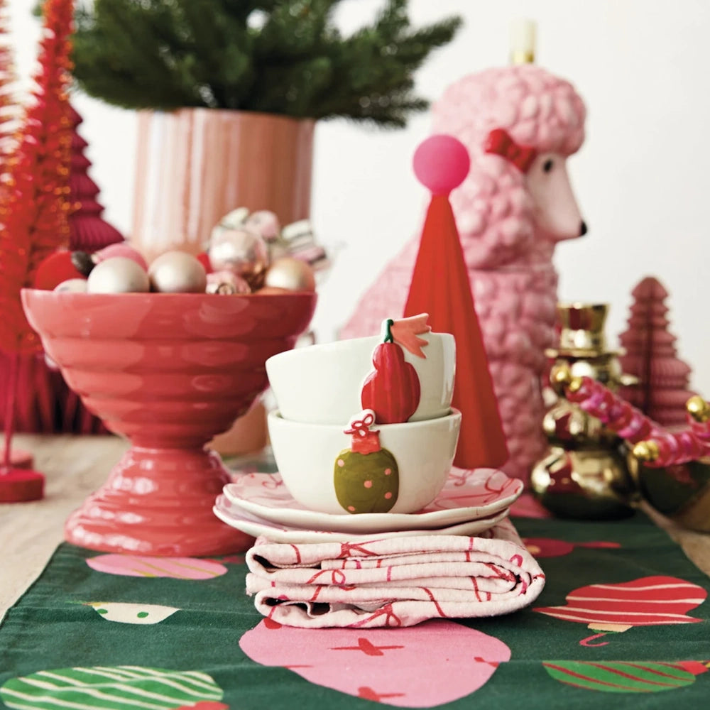 Stacked ornament bowls on pink bow plates atop a patterned runner, surrounded by colorful holiday decor.