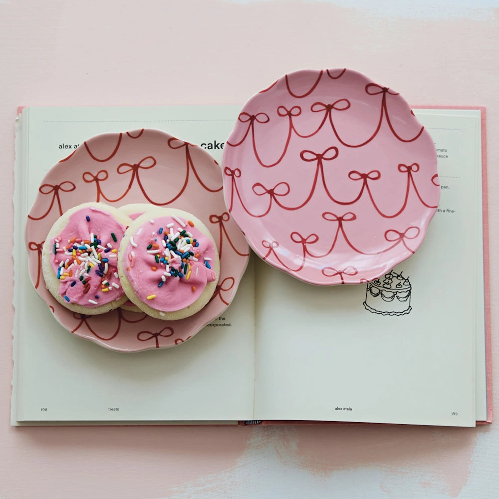 Two pink bow-patterned scalloped plates on an open cookbook, one holding sugar cookies with pink frosting and sprinkles.
