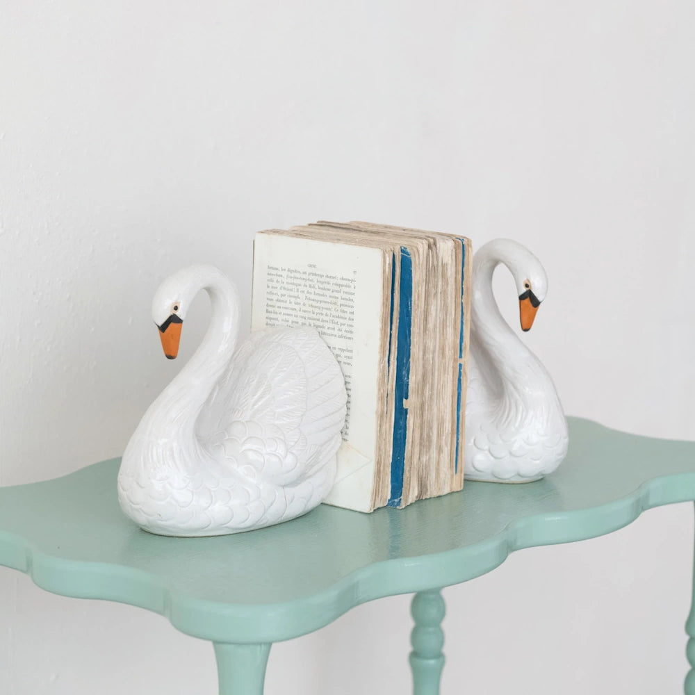 Two white ceramic swan bookends holding vintage books on a teal scalloped console table for elegant home decor.